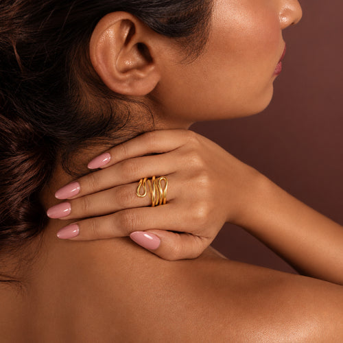 Woman with gold ring on her hand touching her neck against a brown background