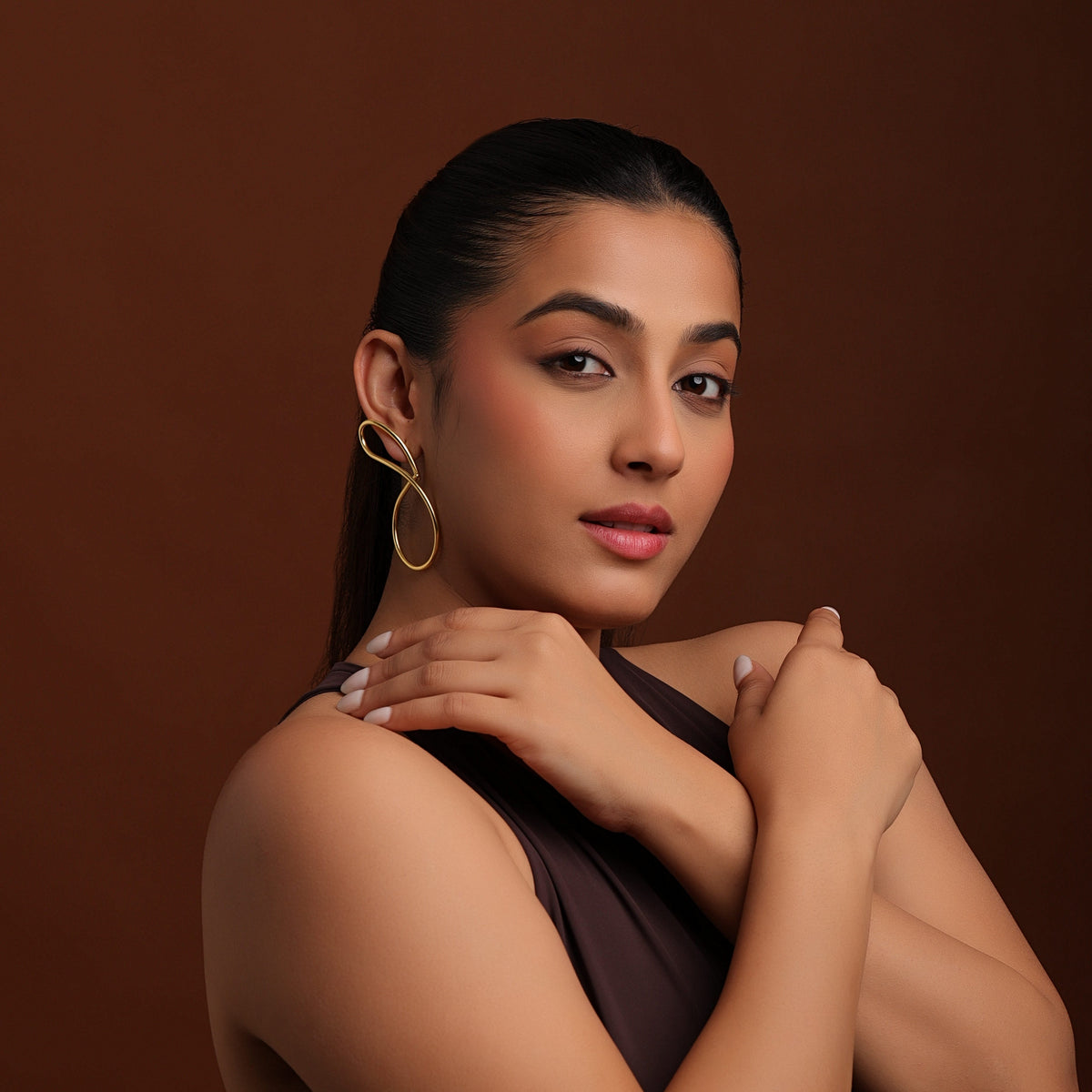 Woman wearing statement gold earrings posing with crossed arms against a brown background