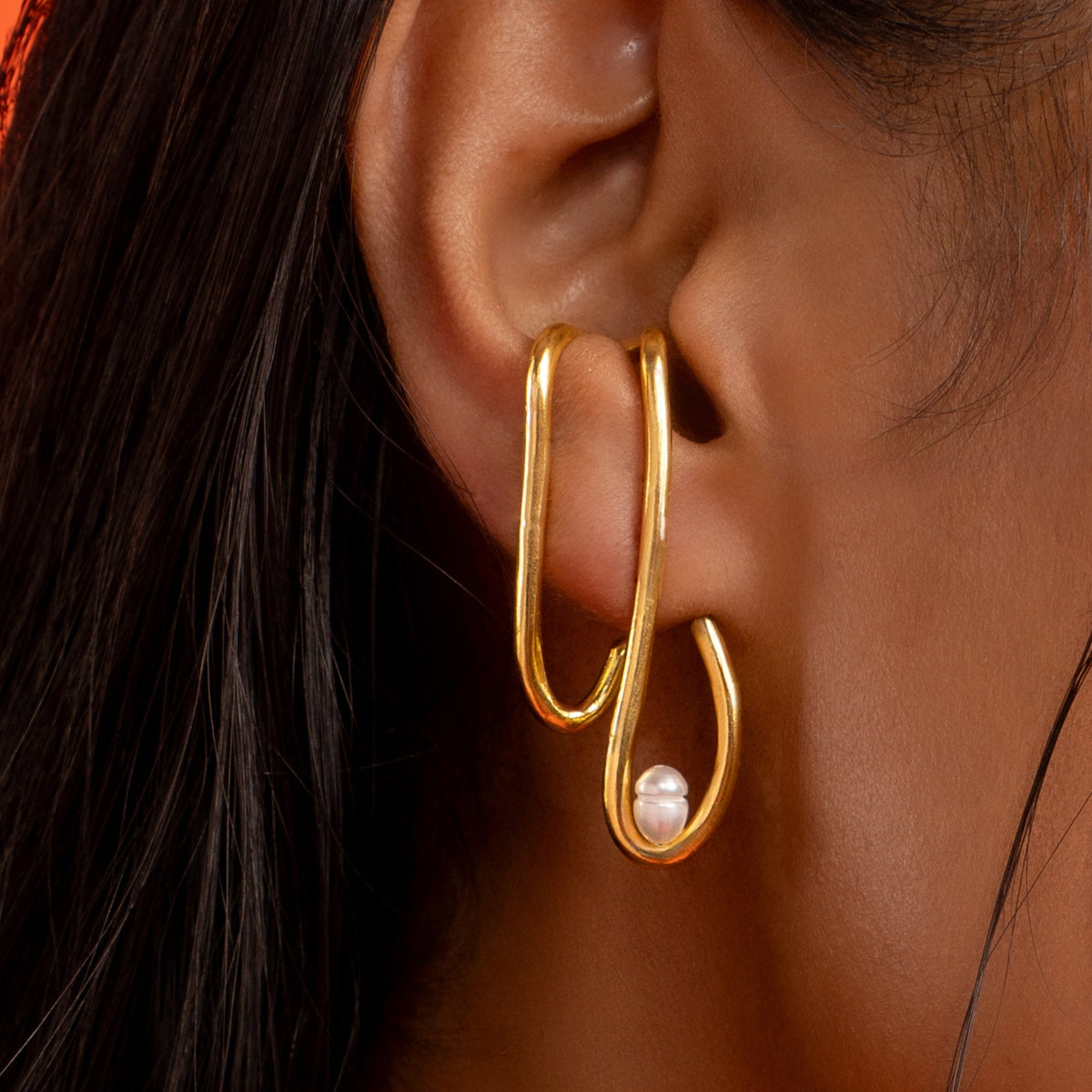 Gold hoop earring with a pearl accent worn by a person against an orange background
