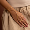 Hand wearing gold arm and hand jewelry against a neutral background