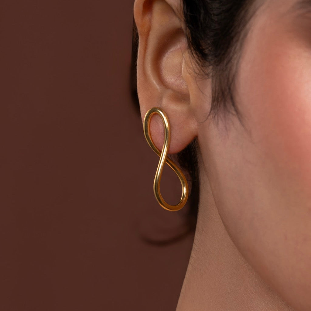 Gold infinity-shaped earring worn by a person against a brown background